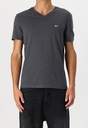 Mężczyzna w gładkiej, ciemnoszarej koszulce typu V-neck z krótkim rękawem, z małym prostokątnym logo na lewej piersi, w połączeniu z czarnymi spodniami.