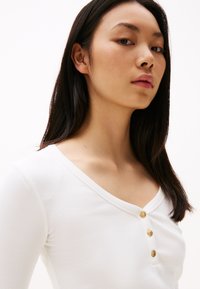 White ribbed long-sleeve top featuring a V-neck and four golden buttons at the neckline. Smooth texture. Simple and minimalistic design.
