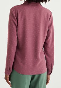 Pitkähihaista pörröruusuista paitaa, jossa on teksturoitu kuvio, hienovaraiset napitusyksityiskohdat ja kaareva helma. Yhdistetty vihreisiin housuihin.