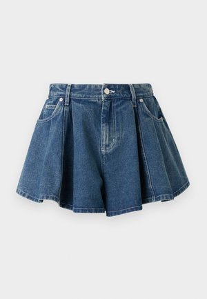 Femeie cu păr lung și întunecat care poartă un tricou alb cu dungi și mâneci scurte, fustă plisată din denim și pantofi Mary Jane roșii închis, cu brațele încrucișate.