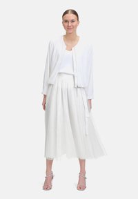 Veste bomber scintillante blanche sur un haut blanc, associée à une jupe plissée blanche fluide. Des sandales argentées à brides complètent le look.
