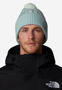 Gorro de punto azul claro con un pompón en la parte superior, que presenta una textura de canalé y un patrón de logo blanco, llevado con una chaqueta negra.