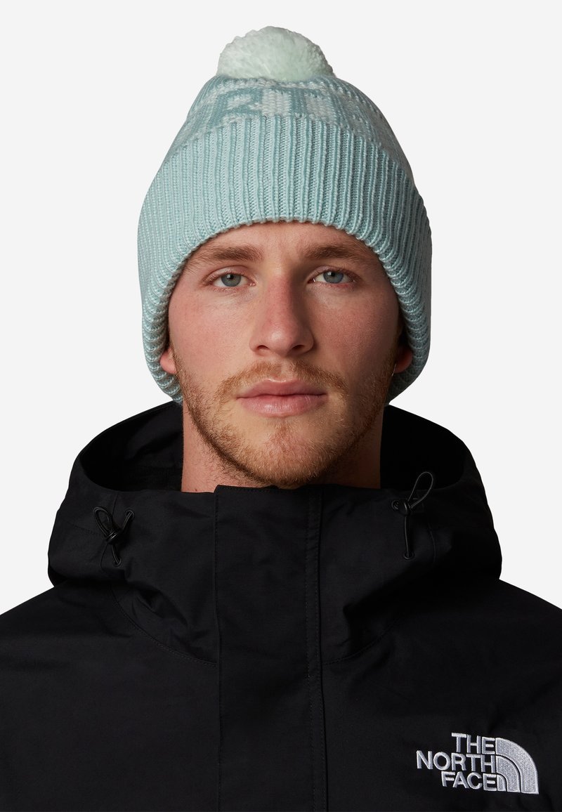 Gorro de punto azul claro con un pompón en la parte superior, que presenta una textura de canalé y un patrón de logo blanco, llevado con una chaqueta negra.