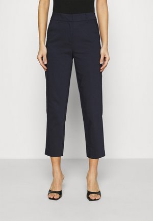 Personne portant un pantalon bleu marine foncé raccourci et ajusté ainsi que des sandales à talons noires à bout ouvert, debout devant un fond clair uni.