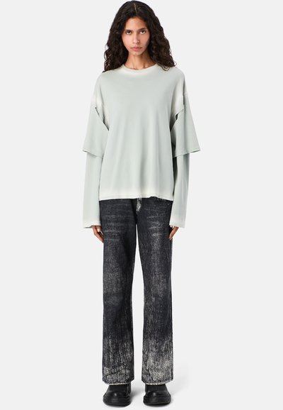 Lichtgrijze oversized top met brede mouwen, gecombineerd met een zwarte jeans met hoge taille en een vervaagde textuur. Zwarte schoenen maken de look compleet.
