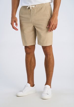 Mężczyzna ubrany w beżowe sztruksowe szorty ze ściągaczem w pasie i białe trampki, stojący na jednolitym tle.