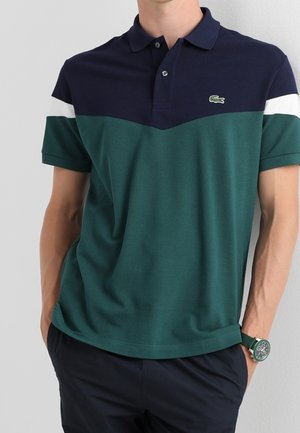 Homme portant un polo à coloris bloqué bleu marine et vert avec une bande blanche et un logo crocodile vert, les mains dans les poches, et une montre verte.