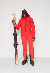 Férfi piros sídzsekiben és nadrágban, fekete síbakancsban és nagy fekete sígoggliban, egy pár sílécet függőlegesen tart.