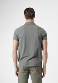 Hombre de cabello oscuro que lleva una camiseta polo gris de manga corta y pantalones verde oliva, de pie de espaldas sobre un fondo liso.