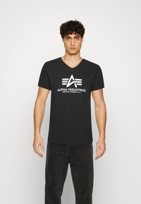 Černé tričko s výstřihem do V vyrobené z bavlny, s bílým logem "ALPHA INDUSTRIES" a třemi horizontálními pruhy vpředu.