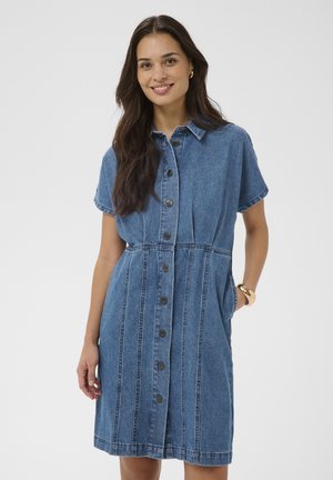 Kvinde med langt mørkt hår iført en kortærmet blå denimkjole med knapper, smilende med den ene hånd i lommen mod en ensfarvet baggrund.