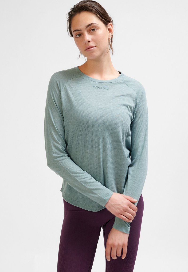 Langärmliges, mintgrünes Sportshirt aus leichtem Stoff mit Rundhalsausschnitt und dezentem Logo auf der Brust, kombiniert mit dunkelvioletten Leggings.