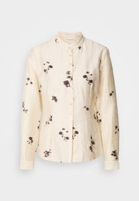 Μπεζ μακρυμάνικη μπλούζα με γιακά μανδαρίνι, κουμπιά μπροστά, διακριτικό μαύρο λουλουδάτο σχέδιο και υφή υφάσματος.