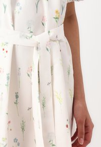 Vestido com padrão floral em tecido leve, apresentando um fundo branco, flores em várias cores pastel e um cinto amarrado na cintura.