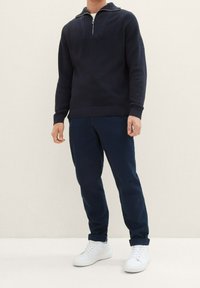 Pullover in maglia blu navy con collo a zip a un quarto, spalle testurizzate e polsini a coste, abbinato a pantaloni blu navy scuri e sneakers bianche.