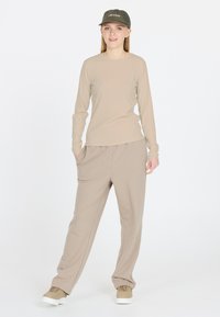 Beige langärmliges Rippoberteil kombiniert mit lockeren beigen Hosen. Dazu eine grüne Kappe mit dem Aufdruck "LES ARCS" und sandfarbene Sneakers. Schlichte, neutrale Farbpalette.