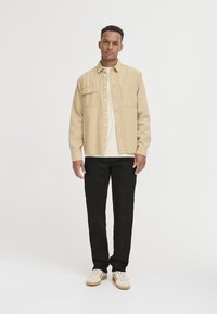 Beige kordfløyels skjortejakke med to frontlommer, over en kremfarget t-skjorte, parret med sorte bukser og hvite joggesko med beige detaljer.