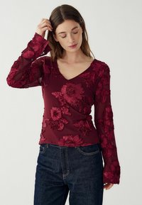 Naine, kandes läbipaistvat burgundilõhku pikka varrukat ja lilledest tikanditega ning tumedatest sinistest teksadest, puutub oma juustesse ja vaatab allapoole.