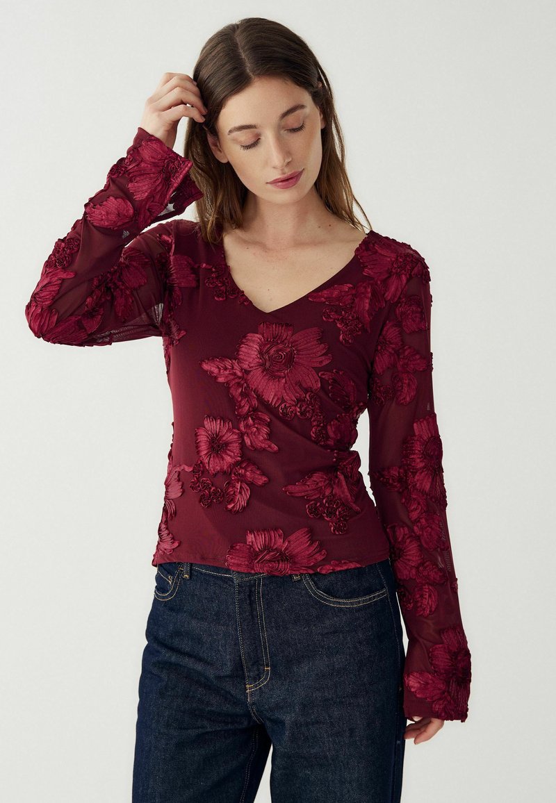 Naine, kandes läbipaistvat burgundilõhku pikka varrukat ja lilledest tikanditega ning tumedatest sinistest teksadest, puutub oma juustesse ja vaatab allapoole.