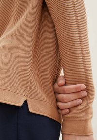 Pull en tissu pêche avec un design côtelé et des fentes latérales à l'ourlet. Une main repose sur la manche, mettant en valeur les détails du matériau.