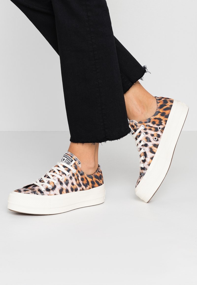 Osoba ubrana w czarne, krótkie spodnie oraz trampki w panterkę z białymi grubymi podeszwami i sznurowadłami na jasnej, gładkiej podłodze.