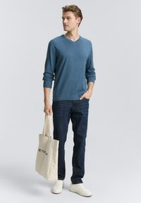 Blauer gestrickter V-Ausschnitt-Pullover mit langen Ärmeln, kombiniert mit dunklen Jeans und weißen Sneakers. Eine beige Tote-Tasche wird in der linken Hand gehalten.