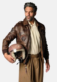 Blouson en cuir marron avec des patchs, t-shirt côtelé crème, pantalon marron et un casque de pilote marron avec un revêtement en fourrure.