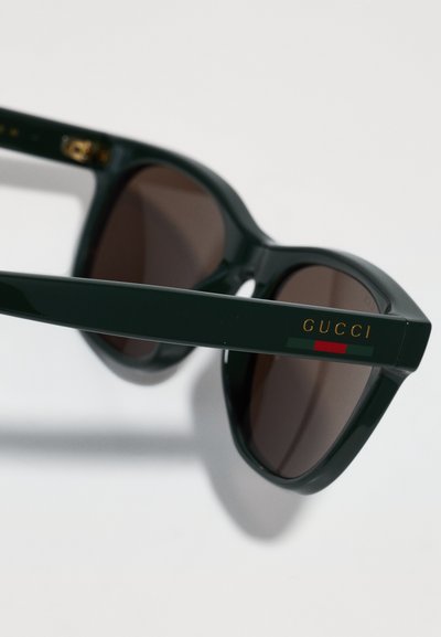 Gucci saulesbrilles ar tumši brūniem lēcu, zaļu plastmasas rāmi un zelta emblēmu uz tempļa, ar sarkaniem un zaļiem akcentiem.