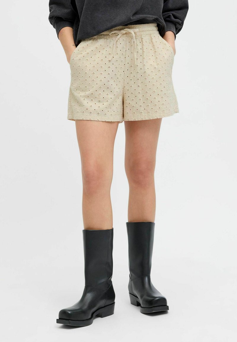 Cremfarbene Shorts mit gesticktem Punkte-Muster, elastischem Bund, Kordelzug und Seitentaschen, kombiniert mit schwarzen kniehohen Stiefeln.