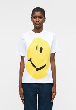 Kvinde iført en hvid t-shirt med stort gult smiley-ansigt, kombineret med mørkeblå jeans, stående mod en ensfarvet baggrund.