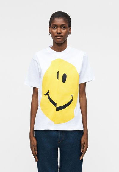 Femme portant un t-shirt blanc avec un grand visage souriant jaune, associé à un jean bleu foncé, debout devant un fond uni.