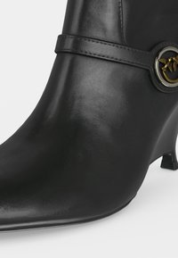 Bottine en cuir noir à petit talon, avec une sangle décorative et un emblème métallique rond avec des oiseaux dorés sur le côté.