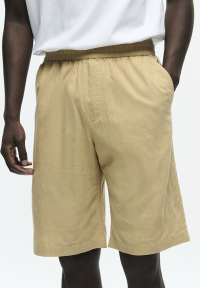 Homme portant un short beige mi-long avec une taille élastique et des poches latérales, associé à un t-shirt blanc.