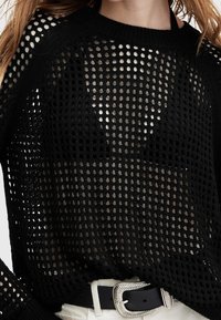 Pull noir à maille lâche porté sur un soutien-gorge noir avec un pantalon blanc et une ceinture noire ornée d'une boucle décorative argentée.