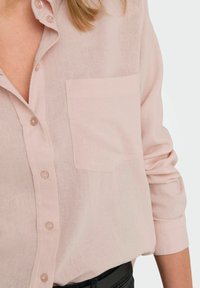 Blusa de botones rosa claro hecha de un tejido ligero, con un bolsillo en el pecho y mangas enrolladas.