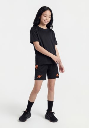 Fiatal lány áll, fekete sportfelszerelésben narancssárga Arsenal és Adidas logókkal, fekete zokniban és fekete sportcipőben.
