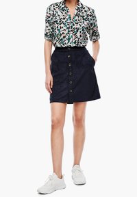 Blusa de estampado floral en colores claros, combinada con una falda en corte A de ante oscura con botones frontales y bolsillos laterales. Zapatillas blancas.