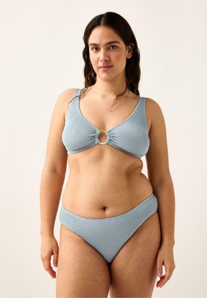 Pitkähiuksinen nainen, jolla on teksturoidusta vaaleansinisestä bikinistä yläosan renkaalla yksityiskohta, seisomassa yksinkertaista vaaleaa taustaa vasten.