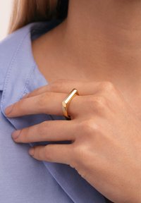 Hand mit hellem Hautton, die einen polierten goldenen geometrischen Ring am Mittelfinger trägt, am Kragen eines hellblauen Hemdes liegt.