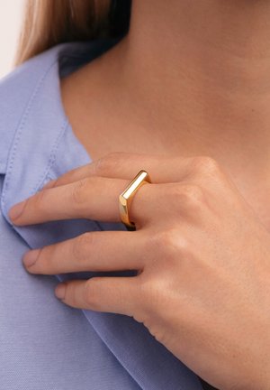Hand mit hellem Hautton, die einen polierten goldenen geometrischen Ring am Mittelfinger trägt, am Kragen eines hellblauen Hemdes liegt.