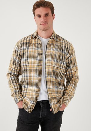 Camisa de cuadros amarilla y negra con cuello, que presenta mangas largas, un patrón de cuadros y un bolsillo en la parte delantera. Se lleva sobre una camiseta blanca.