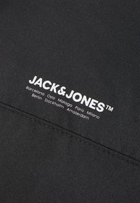 Tessuto nero con texture liscia presenta il logo "JACK & JONES" in bianco, accompagnato dai nomi delle città stampati sotto.