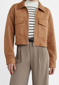Veste en daim beige coupée avec poches avant, portée sur un haut rayé blanc et noir, associée à un pantalon beige plissé taille haute et une ceinture noire.