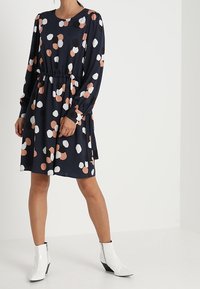 Granatowa sukienka z okrągłym dekoltem i długimi rękawami, wyróżniająca się wzorem w kropki w odcieniach brzoskwini, czerni i bieli; elastyczny pas, długość do kolan.