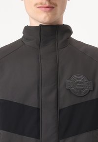 Giacca zip-up nera e grigia con colletto alto, dotata di un distintivo testurizzato sul petto e un design a blocchi di colore con cuciture a contrasto.