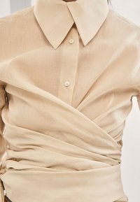 Chemise portefeuille beige en tissu léger, dotée d'un col classique, d'une fermeture à boutons et d'une texture douce avec un design drapé.