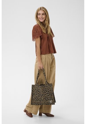 Ženska, oblečena v rjavi zgornji del z čipkasto rokavico in široke bež hlače, drži torbo s tigrastim potiskom, gleda vstran in se smehlja.