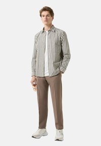 Gestreept overhemd met knopen in beige en donkergrijs, gecombineerd met taupekleurige broek. Model staat en draagt witte sneakers en houdt een bril vast.