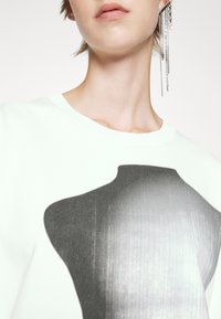 Ljusgrön sweatshirt med en grafik av en docka. Har en ribbad halsringning och en slät textur. Silverörhängen med kedjor är synliga.