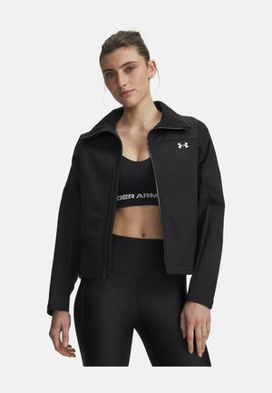 Vrouw in een zwarte jas met rits over een zwarte sportbeha en zwarte leggings, staand tegen een effen lichte achtergrond.